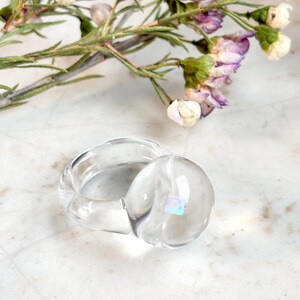 May include: A clear glass ring with a round, iridescent stone set in the center. The ring is on a white surface.