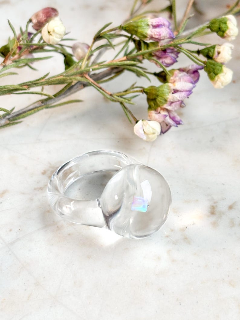 May include: A clear glass ring with a round, iridescent stone set in the center. The ring is on a white surface.