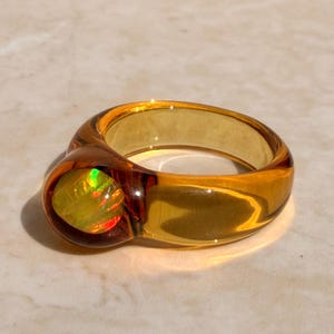 Könnte beinhalten: Ein transparenter, bernsteinfarbener Ring mit einem großen, abgerundeten Edelstein. Der Edelstein hat gelbe, grüne und rote Innenfarben. Der Ring liegt auf einer hellen Oberfläche, wobei Schatten Tiefe verleihen.
