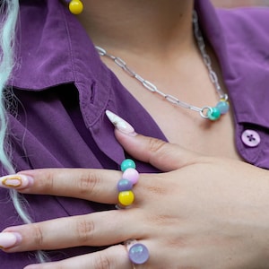 Könnte beinhalten: Ein farbenfrohes Schmuckset mit Halskette, Ohrringen und einem Ring. Die Ohrringe und der Ring haben eine Reihe von bunten Perlen in Grün-, Rosa-, Lila- und Gelbtönen. Die Halskette hat eine silberne Kette mit ein paar bunten Perlen.