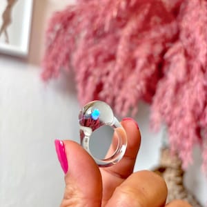 May include: A clear glass ring with a iridescent blue center. The ring is on a finger.