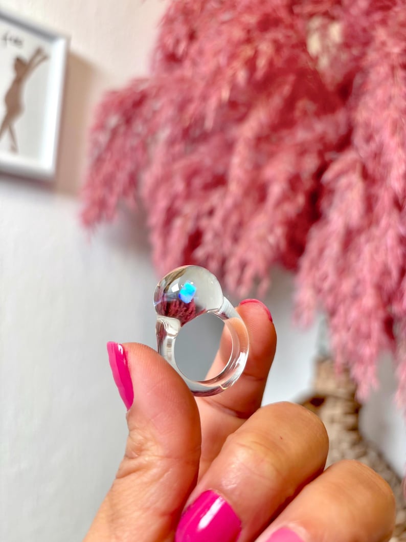 May include: A clear glass ring with a iridescent blue center. The ring is on a finger.