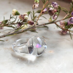 May include: A clear glass ring with a swirl design and a small iridescent pink stone embedded in the center.