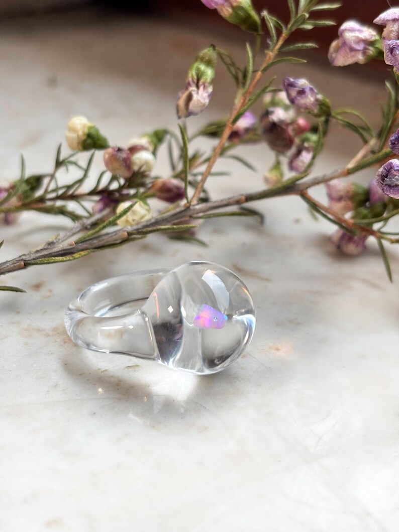 May include: A clear glass ring with a swirl design and a small iridescent pink stone embedded in the center.