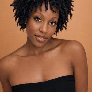 May include: A woman with dark skin and short, dark, curly hair. She is wearing a black strapless top. The background is a solid, warm, orange-brown color. She is looking at the camera with a slight smile.