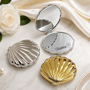 May include: Three shell-shaped compact mirrors in silver and gold finishes. One silver mirror is open, revealing a reflective surface with the name Hanna and a small flower icon engraved on the bottom half. The other two mirrors are closed.