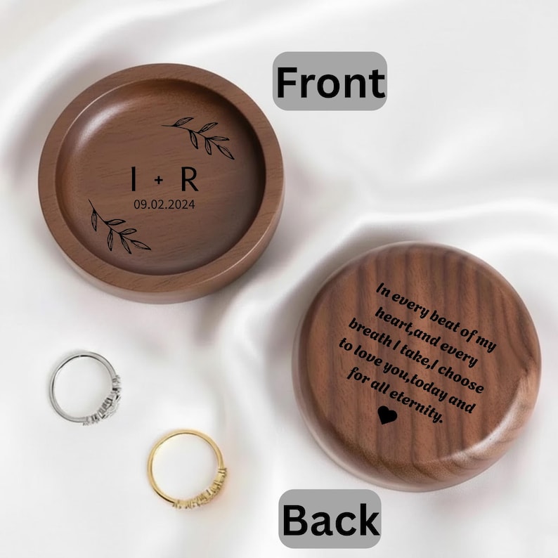 May include: A round, wooden ring holder with the initials "I + R" and the date "09.02.2024" engraved on the front. The back of the holder has a romantic quote and a heart symbol. Two rings, one silver and one gold, are next to the holder.