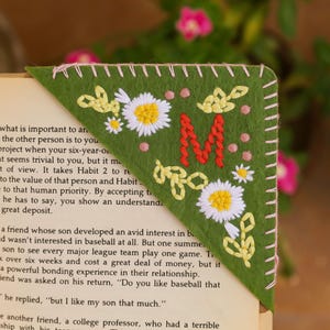 May include: A triangular olive green felt corner bookmark featuring hand-embroidered details including a large red letter M, white and yellow daisy-like flowers, small yellow leaves, and pink dots, finished with light pink whip-stitched edges.