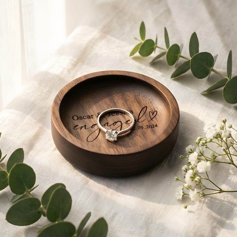 Handcrafted walnut wood ring dish personalized with names and engagement date, holding a diamond engagement ring, styled with eucalyptus leaves and baby's breath on a linen background.