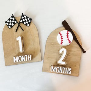 May include: Wooden milestone markers for baby photos. One marker features checkered flags and the number 1 with the word "MONTH". The other has a baseball, bat, and the number 2 with the word "MONTHS".