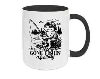 May include: White ceramic mug with a black interior and handle. The mug features a black and white illustration of a frog fishing with the text "GONE FISHIN' Mentally". The design includes a frog, fishing rod, lake, and trees.