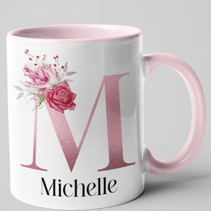 Può includere: Tazza in ceramica bianca con interno e manico rosa. La tazza presenta una grande lettera "M" rosa con il nome "Michelle" sotto. Una composizione floreale con rose rosa e rosse e altri fiori decora il lato sinistro della lettera.
