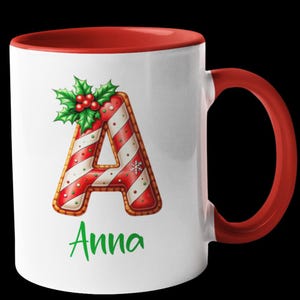 Könnte beinhalten: Weiße Keramik-Tasse mit rotem Innenraum und Henkel. Die Tasse zeigt einen Lebkuchen-Buchstaben "A", verziert mit rot-weißen Streifen, einer Schneeflocke und Stechpalme. Der Name "Anna" ist grün gedruckt.