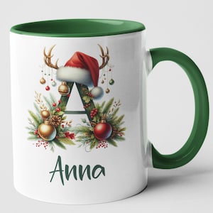 May include: White ceramic mug with a green handle and interior. The mug features a Christmas-themed design with the letter "A" decorated with a Santa hat, antlers, ornaments, and the name "Anna".