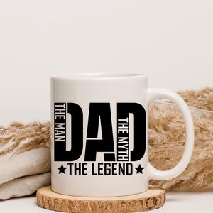 Pode incluir: Caneca de cerâmica branca com o texto "DAD THE MAN THE MYTH THE LEGEND" em letras pretas em negrito. A caneca está sobre um descanso de madeira. O fundo é neutro com plantas secas.