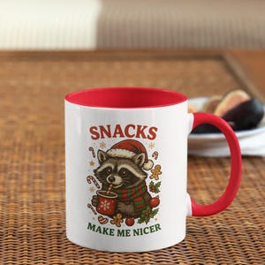 Könnte beinhalten: Weiße Keramiktasse mit rotem Innenraum und Henkel. Die Tasse zeigt einen Waschbären mit Weihnachtsmütze und Schal, der eine Tasse hält. Auf der Tasse steht "SNACKS MAKE ME NICER" mit Zuckerstangen und Lebkuchenmännern.