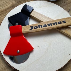 Puede incluir: Dos cortadores de pizza de madera con forma de hacha. Uno es rojo con una cuchilla plateada y el otro es negro con un mango de madera. El cortador rojo tiene el nombre "Johannes" escrito en el mango.