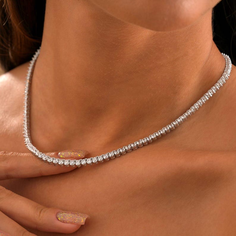 May include: A delicate, silver-toned necklace with numerous small, round, clear stones. The necklace is worn around the neck, and the stones sparkle. The image is a close-up, highlighting the necklace's design and the skin tone of the person wearing it.