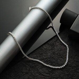May include: A delicate silver necklace with numerous small, round, clear gemstones. The necklace is displayed on a textured silver cylinder and a black, crinkled surface, creating a contrast of textures and colors. The necklace is a jewelry item.