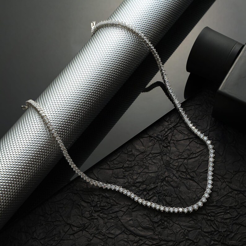May include: A delicate silver necklace with numerous small, round, clear gemstones. The necklace is displayed on a textured silver cylinder and a black, crinkled surface, creating a contrast of textures and colors. The necklace is a jewelry item.