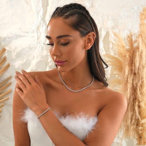 May include: A silver necklace and bracelet set. The jewelry features a delicate design with small, evenly spaced stones. The pieces are worn on a person with a white top and a neutral background.