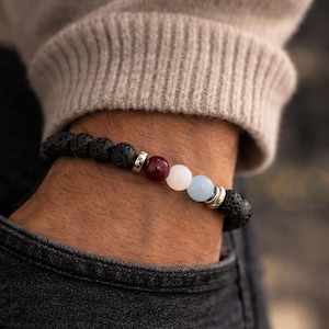 May include: A black lava stone bead bracelet with a mix of colorful gemstone beads. The bracelet features a dark red, white, and light blue beads, separated by silver-colored metal spacers. The bracelet is worn on a wrist.