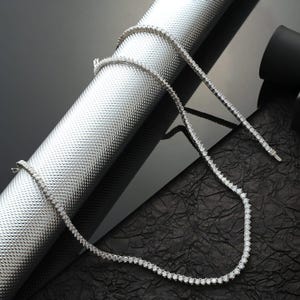 May include: A delicate, silver-toned necklace with numerous small, round, clear gemstones. The necklace is displayed on a textured, metallic silver surface and a black, crinkled surface, creating a contrast. The necklace is a tennis style.
