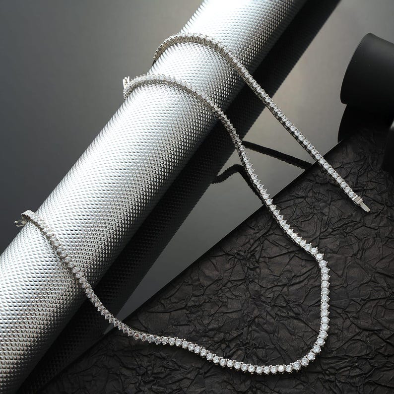 May include: A delicate, silver-toned necklace with numerous small, round, clear gemstones. The necklace is displayed on a textured, metallic silver surface and a black, crinkled surface, creating a contrast. The necklace is a tennis style.