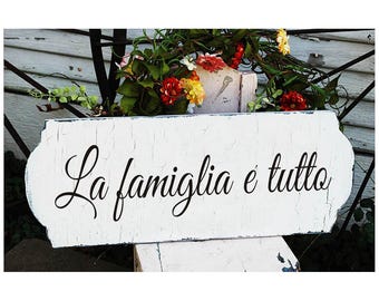 May include: White wooden sign with distressed paint and rounded edges. The sign features black cursive lettering that reads "La famiglia è tutto".
