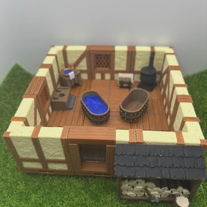 May include: A miniature diorama of a medieval bathhouse. The structure features a wooden frame with cream-colored walls and brown beams. Inside are two wooden tubs, one filled with blue water. A small wood storage area is attached to the side.