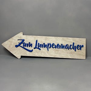 Puede incluir: Un letrero de flecha de madera con el texto "Zum Lumpenmacher" en letras azules.