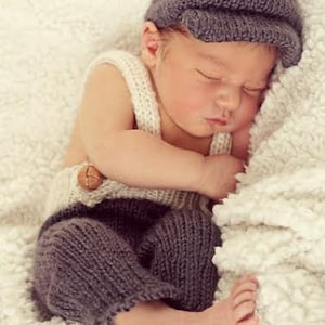 May include: A sleeping baby wearing a gray knitted hat and matching gray knitted overalls with a brown button. The baby is lying on a white fluffy blanket.