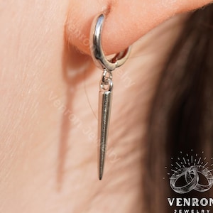May include: Silver hoop earrings with a dangling silver spike. The earrings are worn in a person's ear.