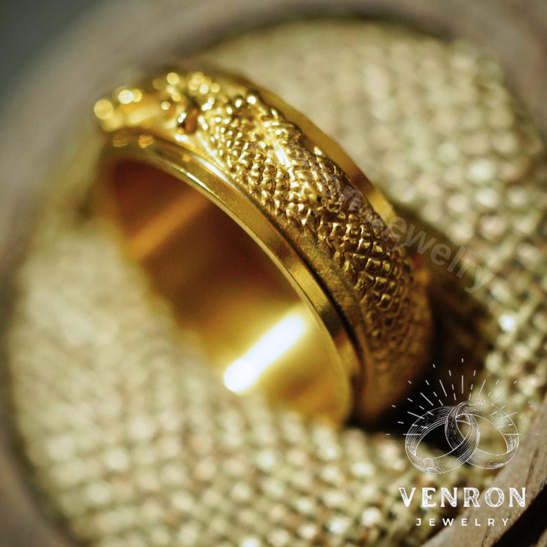 May include: A gold ring with a textured dragon design. The ring is shown on a textured fabric background.