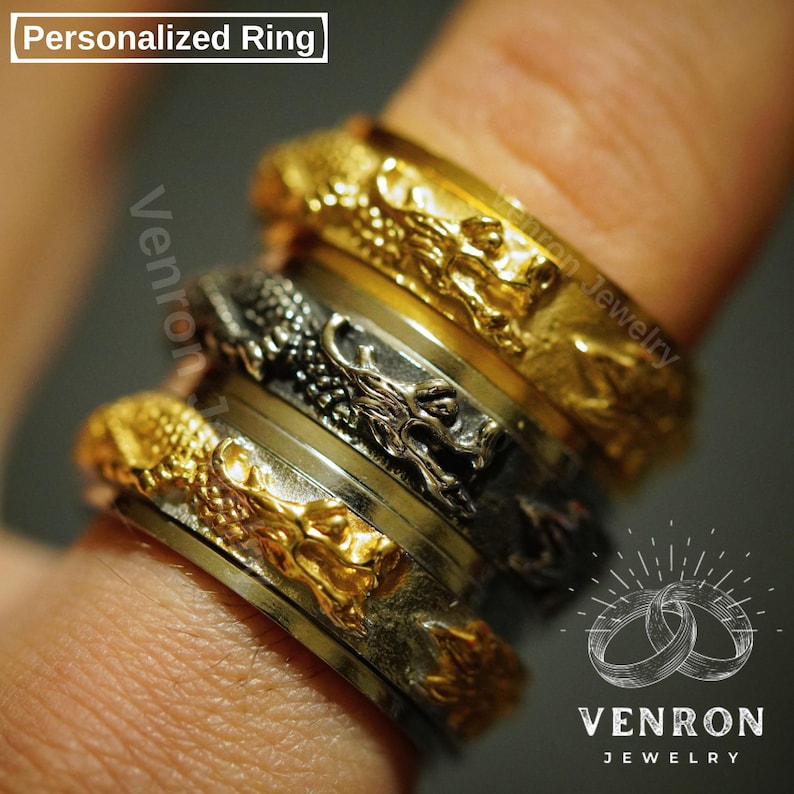 May include: A set of four stackable rings featuring a dragon design. The rings are made of gold and silver metal. The top ring is gold with a dragon design, the second ring is silver with a dragon design, the third ring is silver, and the bottom ring is gold with a dragon design. The rings are stacked on a finger.