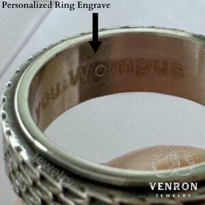 May include: Close-up of a silver ring with a detailed design. The inside of the ring is engraved with the words "You Wompus".