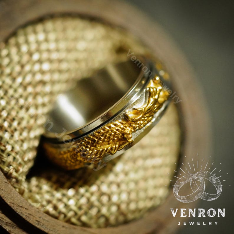 May include: A gold and silver spinning ring with a dragon design. The ring is on a brown and beige textured surface. The text "VENRON JEWELRY" is visible in the lower right corner of the image.