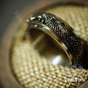 May include: A silver ring with a detailed dragon design. The dragon is in a dark gray color with gold accents. The ring is on a brown fabric background.