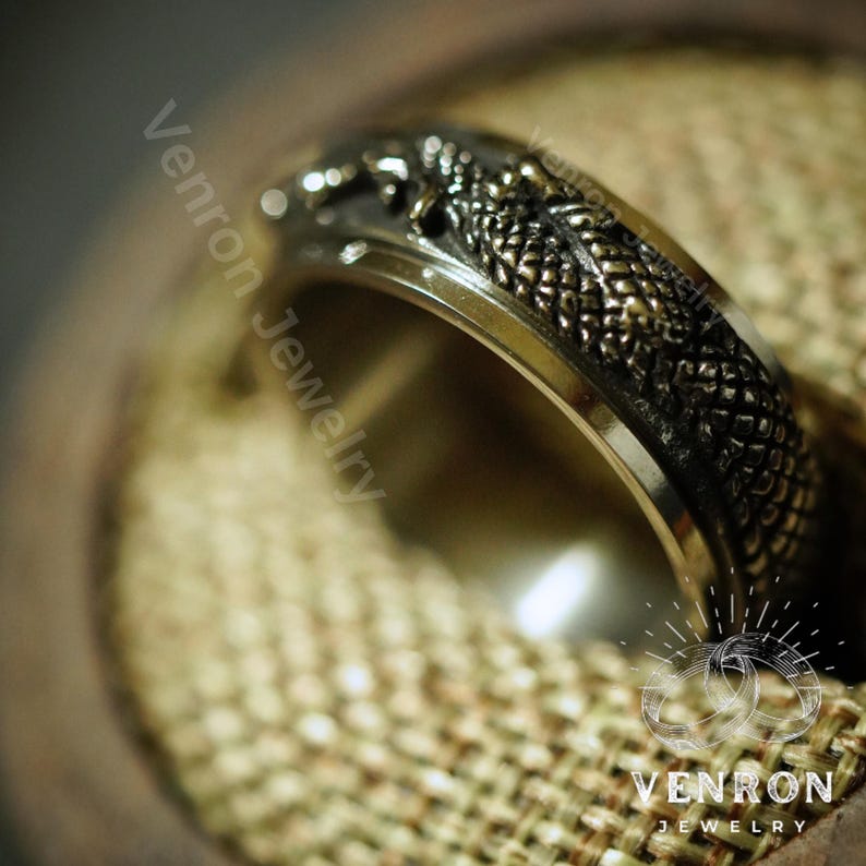 May include: A silver ring with a detailed dragon design. The dragon is in a dark gray color with gold accents. The ring is on a brown fabric background.