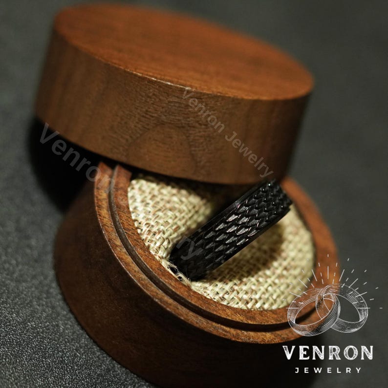 May include: A black ring with a textured pattern sits inside a wooden ring box. The box is made of dark wood and has a light brown fabric lining. The box is labelled "Venron Jewelry".