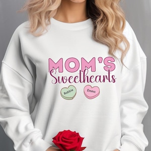 Può includere: Felpa bianca con il testo "MOM'S Sweethearts" in lettere rosa. Due cuori di caramelle rosa con i nomi "Robbie" ed "Emma" sono sotto il testo.