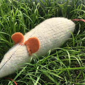 May include: A white knitted mouse toy with orange felt ears lying in green grass.