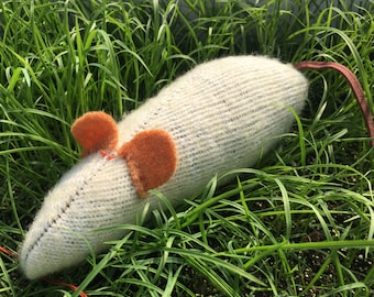 May include: A white knitted mouse toy with orange felt ears lying in green grass.