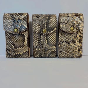 May include: Three rectangular vertical cases featuring a textured snakeskin pattern in shades of brown, black, and cream. Each case has a flap closure secured with a single brass-colored metal snap button centered on the front of the flap.