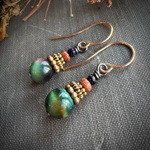May include: A pair of handcrafted earrings featuring green and gold cat's eye beads. The earrings have gold-colored ear wires and are accented with small gold, orange, and dark blue beads. The earrings are displayed on a textured gray surface.