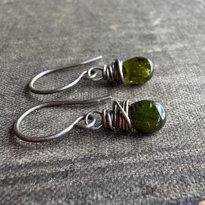 May include: A pair of handcrafted earrings featuring olive-green gemstones wrapped in silver-colored wire. The earrings have a simple hook design. The gemstones are teardrop-shaped and have a polished, glossy finish. The earrings are displayed on a textured, gray surface.