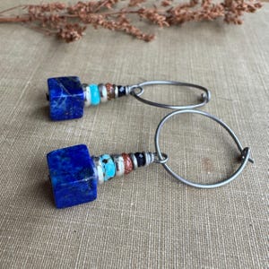 May include: A pair of handcrafted earrings featuring square, deep blue lapis lazuli stones. The earrings have a silver-tone metal wire design with turquoise, silver, and black beads. The earrings are a unique accessory.