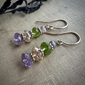 May include: Gold-coloured earrings with faceted gemstones. The earrings feature amethyst, champagne-coloured, and green gemstones, separated by small gold beads. The earrings have a simple hook design.