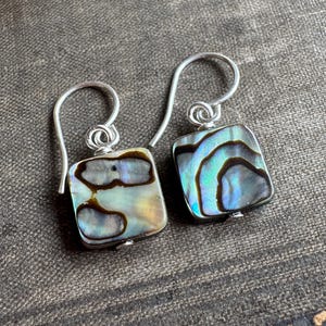 May include: A pair of square-shaped earrings featuring iridescent abalone shell inlays with swirling patterns of blue, green, and brown. The earrings are suspended from silver-toned metal hooks with a simple loop design at the top of each square.