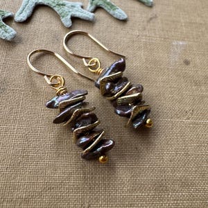 May include: A pair of gold earrings with a cluster of iridescent gray baroque pearls. The pearls are stacked vertically and have a slightly irregular shape. The earrings are made with gold wire and have a simple hook closure.